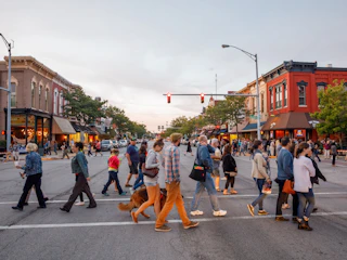 People crossing a busy street in Elkhart County with storefronts and buildings lining both sides of the road.