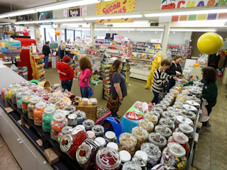 Interior of a candy store with rows of glass jars filled with sweets and shoppers browsing aisles of nostalgic treats.
