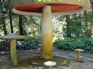 Large concrete mushroom sculpture surrounded by smaller mushroom-shaped seats in a shaded forest garden.