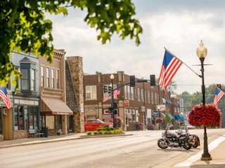 Quiet downtown Wakarusa street with American flags, vintage buildings, hanging flower baskets, and motorcycles under a partly cloudy sky.