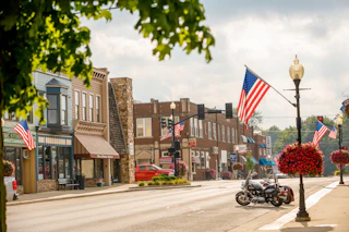 Quiet downtown Wakarusa street with American flags, vintage buildings, hanging flower baskets, and motorcycles under a partly cloudy sky.